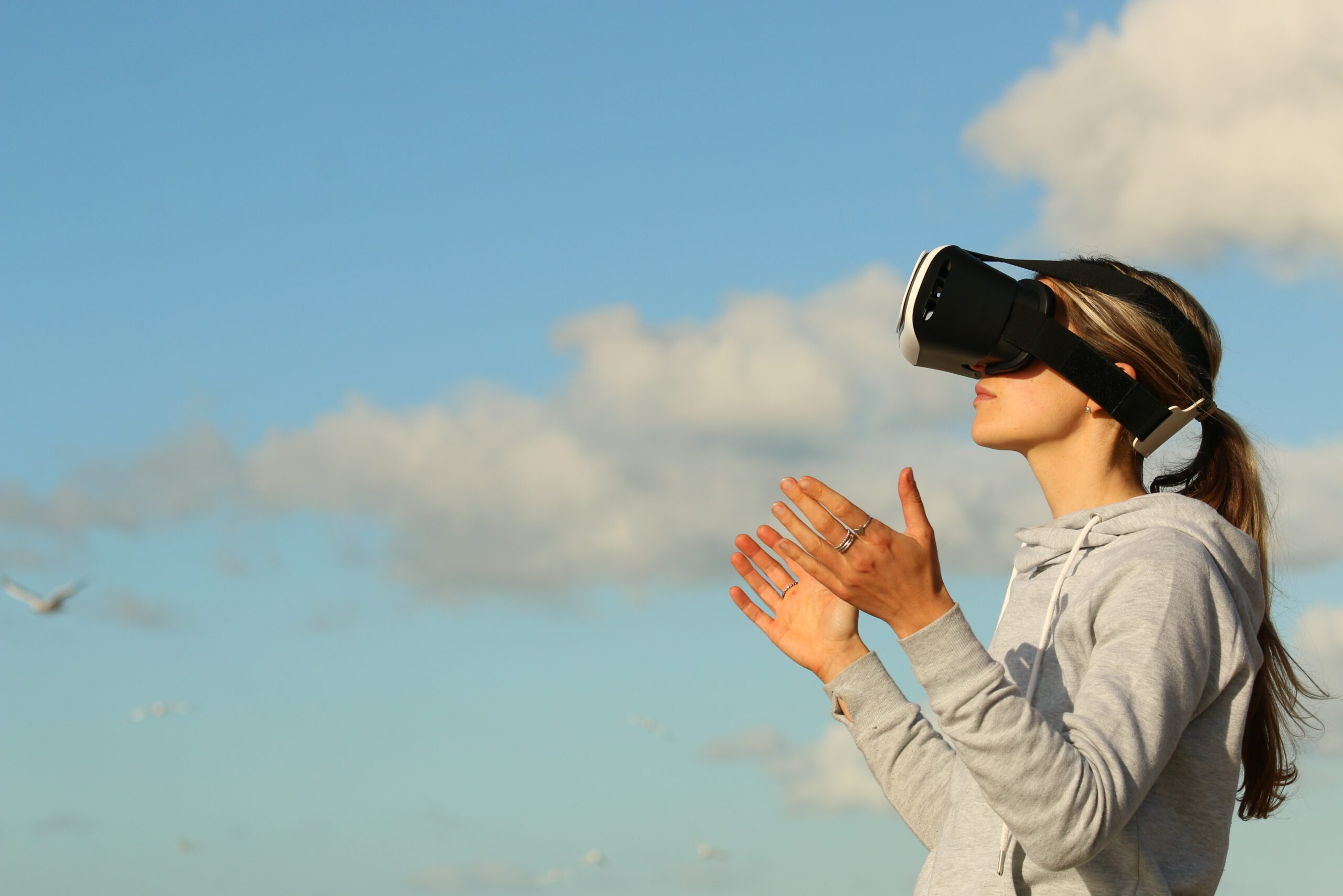 图片展示一位女士戴着虚拟现实头盔，伸手触摸，仿佛在体验虚拟世界，背景是晴朗的天空和飞鸟。